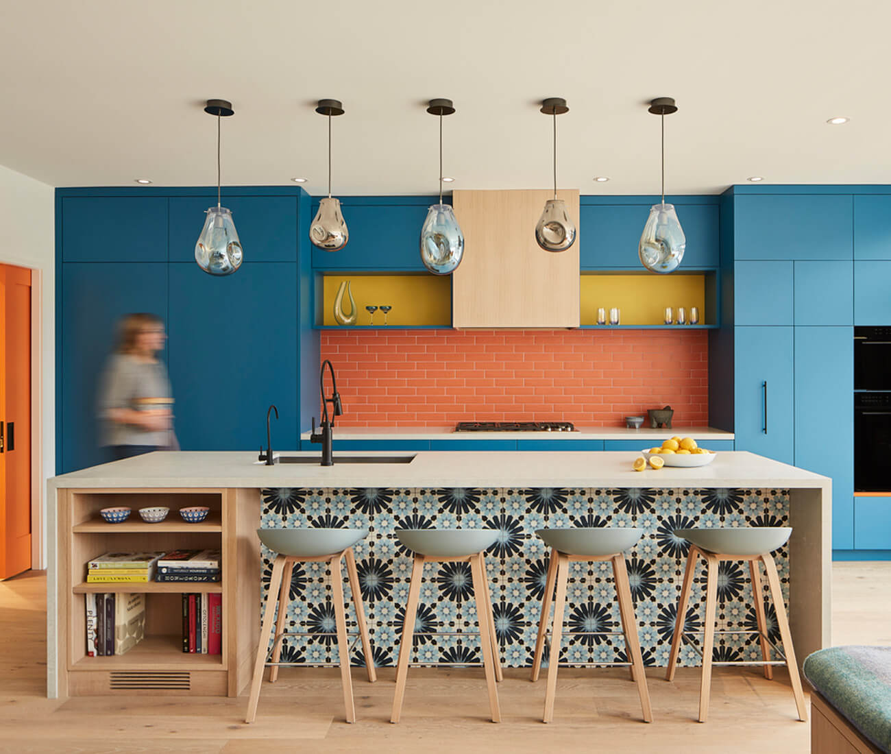 The island is clad in quartz from HanStone and backed by ocean blue millwork, orange subway tile, splashes of yellow and orange accents. Neutral elements, including wood shelving, black fixtures and Hay barstools, lend some restraint. The pendants are by AM Studio.